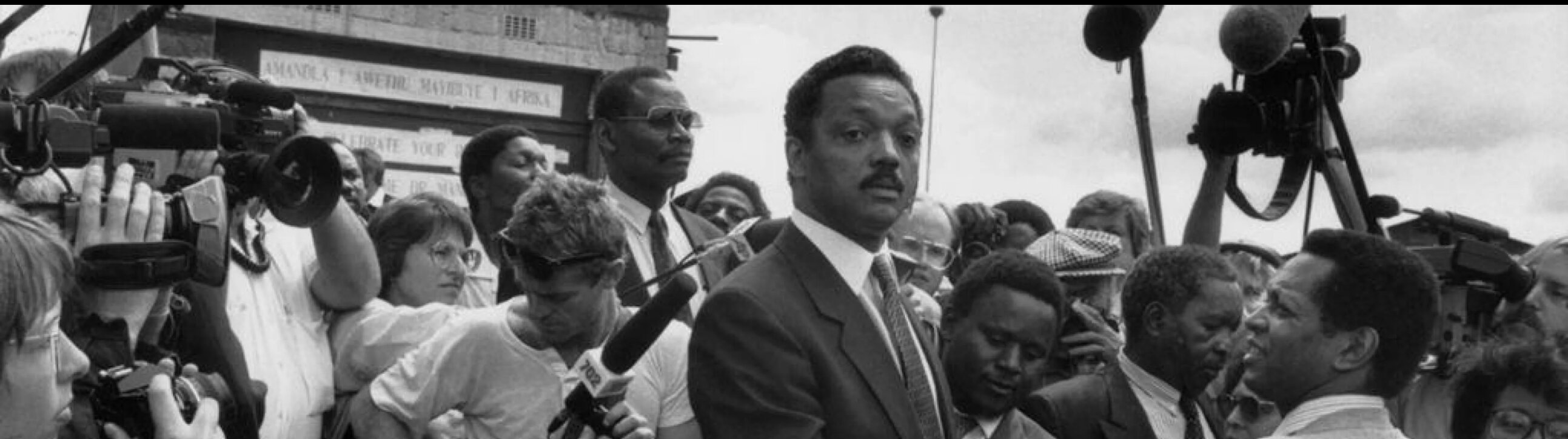 Detail of a black and white photograph by Chris Steele-Perkins depicting American civil rights activist Jesse Jackson during a 1990 visit to Soweto, South Africa. Jackson is surrounded by a dense press scrum of journalists, cameras, and microphones against a backdrop of anti-apartheid signage.